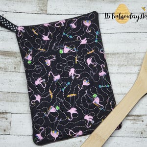 May include: Black pot holder with pink flamingos and a black and white polka dot ribbon handle. The pot holder is on a wooden surface with a wooden spoon.