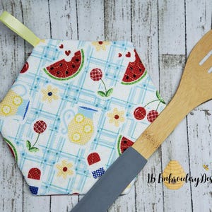 May include: A hexagonal pot holder with a white background and a blue and white checkered pattern. The pot holder features a variety of summer-themed designs, including watermelons, cherries, lemons, and flowers. The pot holder has a yellow loop for hanging.