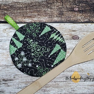 May include: A round, black and green quilted hot pad with a green ribbon loop. The hot pad features a design of green Christmas trees and white snowflakes on a black background. The hot pad is sitting on a wooden surface next to a wooden spoon.