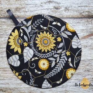 Pu&ograve; includere: Presa pentola rotonda nera con un design floreale che presenta girasoli, foglie e api su uno schema a nido d'ape. La presa pentola ha un anello di sospensione nero.