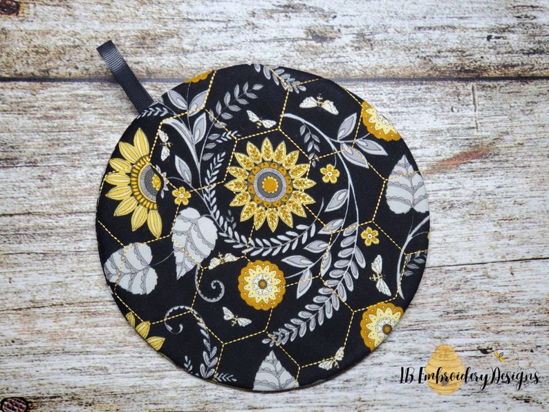 Pu&ograve; includere: Presa pentola rotonda nera con un design floreale che presenta girasoli, foglie e api su uno schema a nido d'ape. La presa pentola ha un anello di sospensione nero.