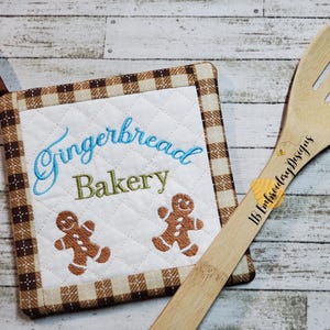 May include: A brown and tan quilted pot holder with a white background. The pot holder has a brown and tan checkered border. The text "Gingerbread Bakery" is embroidered in blue and green on the white background. Two gingerbread men are embroidered on the pot holder in brown.