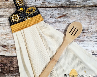 May include: A cream-coloured waffle weave kitchen towel with a black and gold Boston Bruins logo design at the top. A wooden slotted spoon rests on the towel. The towel has a loop for hanging.