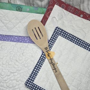 Könnte beinhalten: Ein hölzerner Schöpflöffel mit dem Text "1B Embroidery Designs" liegt auf einer Sammlung von gesteppten Topflappen. Die Topflappen haben verschiedene gemusterte Ränder und weiße, gesteppte Zentren. Die Ränder umfassen grüne Blumen, lila Punkte, rote Paisley-Muster und blaues Vichy-Karo.