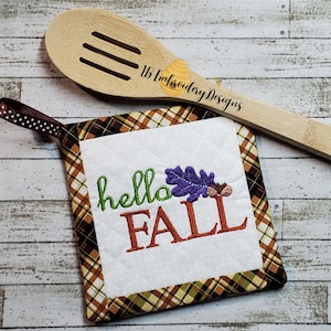 May include: A brown and tan plaid pot holder with a white square in the center. The white square has the text "hello fall" in green and orange with a purple and yellow leaf and acorns.