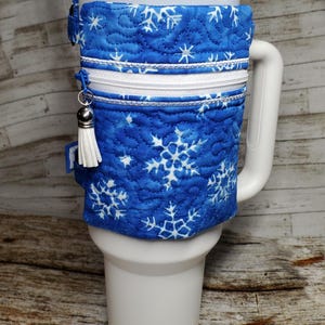 May include: A blue quilted cup holder with a white snowflake pattern and a zipped pocket. The holder is on a white travel mug with a straw.