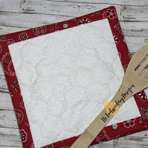 Könnte beinhalten: Ein roter und weißer gesteppter Topflappen mit einem Bandana-Muster am Rand. Ein Holzlöffel mit dem Text "1B Embroidery Designs" liegt auf dem Topflappen.