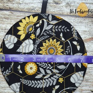 Pu&ograve; includere: Un sottopentola in tessuto nero con un motivo a nido d'ape e un design a girasole. Il sottopentola ha un diametro di 17,8 cm.