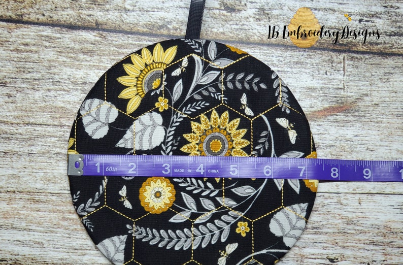 Pu&ograve; includere: Un sottopentola in tessuto nero con un motivo a nido d'ape e un design a girasole. Il sottopentola ha un diametro di 17,8 cm.