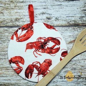May include: Round white quilted hot pad with a red lobster print. The hot pad has a red hanging loop. A wooden spoon is lying on the table next to the hot pad.