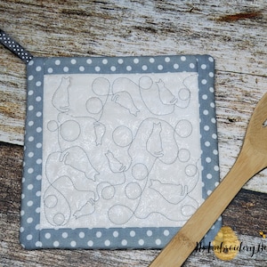 May include: A gray and white quilted hot pad with a design of cats and circles. The hot pad has a gray polka dot border and a black and white ribbon loop.