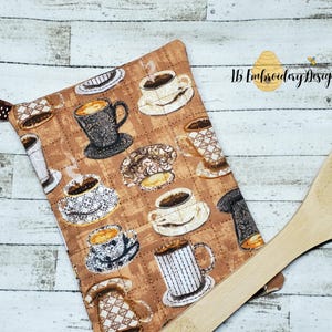 May include: A brown quilted hot pad with a coffee cup design. The hot pad has a loop for hanging. The hot pad is next to a wooden spoon.