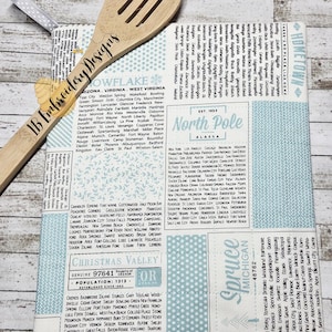 May include: A rectangular fabric potholder featuring a light blue and white newspaper-style print with lists of city names, the text North Pole, Christmas Valley, and Spruce, paired with a wooden slotted spoon branded with IB Embroidery Designs.