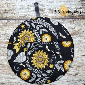 Pu&ograve; includere: Presa pentola rotonda nera con un motivo floreale con girasoli gialli, foglie grigie e piccole api. La presa pentola ha un anello di nastro nero per appenderla.