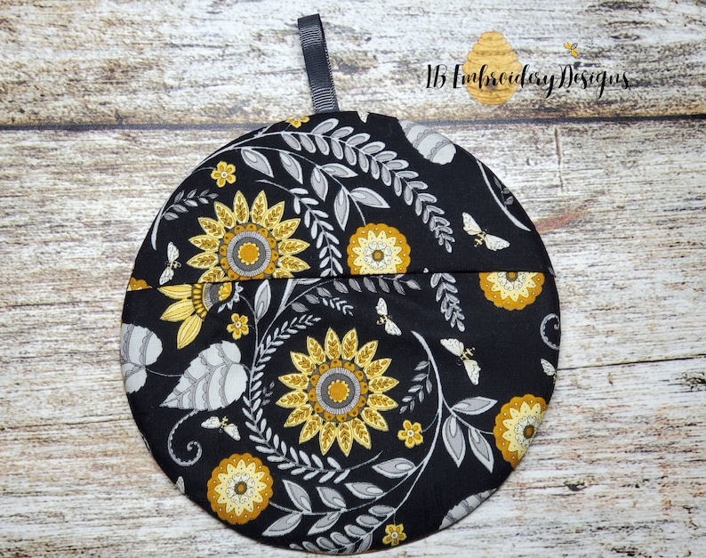 Pu&ograve; includere: Presa pentola rotonda nera con un motivo floreale con girasoli gialli, foglie grigie e piccole api. La presa pentola ha un anello di nastro nero per appenderla.