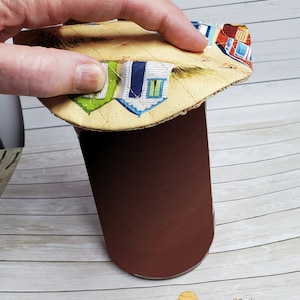 May include: A yellow and blue fabric lid with a beach house pattern. The lid is being placed on top of a brown glass jar.