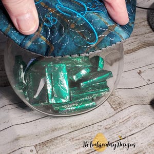 May include: A glass jar filled with individually wrapped green mint sweets. The jar has a fabric lid with the words "So Free" embroidered in blue thread.