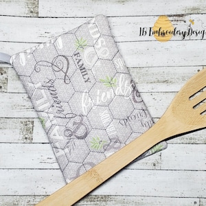 May include: A gray and white quilted hot pad with a chicken wire pattern and the words "Family & Friends" and "Grow Old With Those Who Make You Smile". The hot pad has a loop for hanging. A wooden spoon is lying on top of the hot pad.