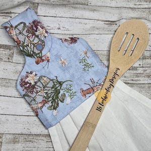 Könnte beinhalten: Ein hellblaues Küchentuch mit einem Meeresleben-Muster, das Hummer, Seesterne und Fischernetze zeigt. Ein hölzerner Schlitzlöffel mit dem Text "IB Embroidery Designs" liegt auf dem Handtuch. Das Handtuch hängt auf einer weißen Oberfläche.