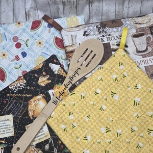 Könnte beinhalten: Eine Sammlung von gesteppten Topflappen mit verschiedenen Mustern, darunter Obst-, Bienen- und Kaffeemotive. Ein hölzerner Schöpflöffel mit "16 Embroidery Designs" befindet sich in der Mitte. Die Topflappen haben Schlaufen zum Aufhängen.