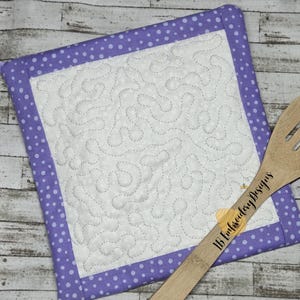 May include: Purple quilted hot pad with white polka dots and a white quilted design. The hot pad has a loop handle and is paired with a wooden spoon with the text "JB Embroidery Designs".