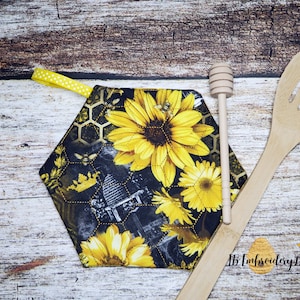 Könnte beinhalten: Sechseckiger Topflappen mit schwarzem Hintergrund und einem Muster aus gelben Sonnenblumen, Bienen und Honigwaben. Der Topflappen hat eine gelbe Schleife zum Aufhängen.