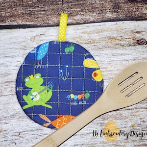 May include: Round, navy blue quilted potholder with a whimsical frog playing a banjo, along with other cartoon creatures. The potholder has a yellow hanging loop and is approximately 7 inches in diameter. The text "Wiggle Ville" is visible on the potholder.