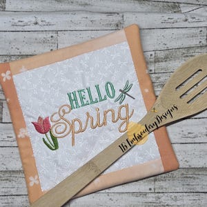 May include: A white pot holder with a peach border features embroidered text that says "Hello Spring" with a stylized tulip and dragonfly. A wooden spoon with the text "BB Embroidery Designs" is in the foreground.