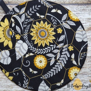 Pu&ograve; includere: Sottopentola rotondo in tessuto nero con un motivo a nido d'ape e un design floreale in giallo e grigio. Il design include girasoli, foglie e api.