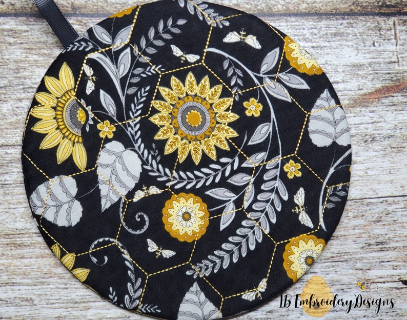 Pu&ograve; includere: Sottopentola rotondo in tessuto nero con un motivo a nido d'ape e un design floreale in giallo e grigio. Il design include girasoli, foglie e api.