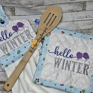 Può includere: Un set di due presine da cucina con il testo "hello winter" ricamato su di esse. Le presine sono blu e bianche con un design trapuntato. È inclusa anche una paletta di legno con il testo "1B Embroidery Designs".