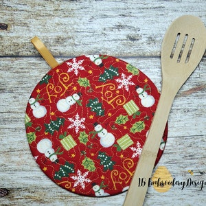 May include: A circular, red Christmas hot pad with a gold ribbon loop. The pad displays a pattern of snowmen, Christmas trees, snowflakes, and gifts. A wooden slotted spoon is placed beside the pad.