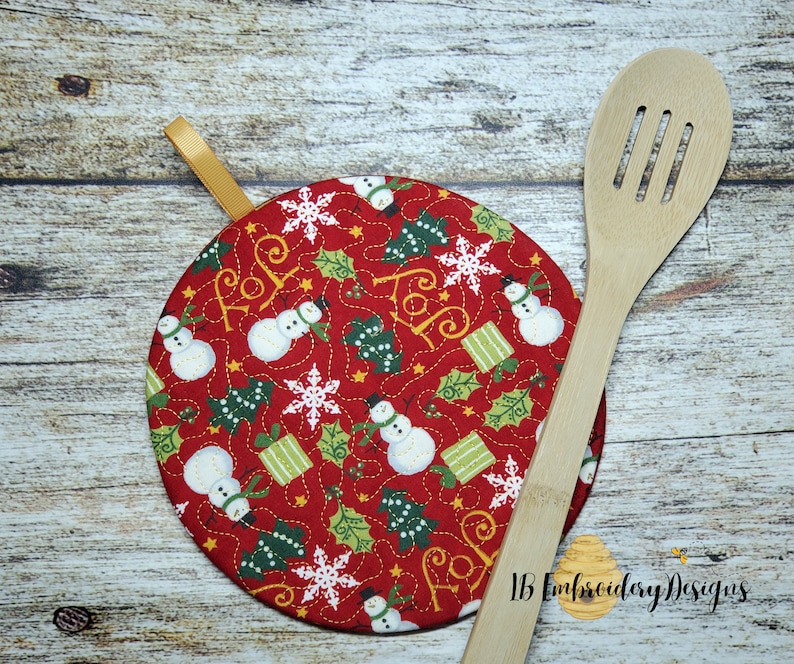 K&ouml;nnte beinhalten: Ein runder, roter weihnachtlicher Topflappen mit einer goldenen Schleife. Das Pad zeigt ein Muster aus Schneem&auml;nnern, Weihnachtsb&auml;umen, Schneeflocken und Geschenken. Ein h&ouml;lzerner Sch&ouml;pfl&ouml;ffel liegt daneben.