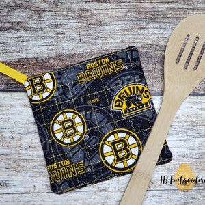 May include: A black and yellow pot holder with the Boston Bruins logo. The pot holder has a yellow loop for hanging. The pot holder is on a wooden surface with a wooden spoon.