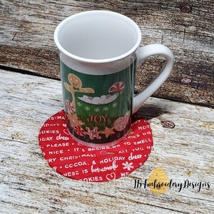 Pode incluir: Copo de tecido de Natal vermelho e branco com o texto "It's beginning to smell like Christmas! All you need is nice Christmas cheer.  Cookies & homemade cocoa.  We  JOY" Uma caneca branca com fundo verde e um desenho de homem de gengibre está sentada em cima do copo.