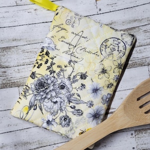 May include: A yellow and white quilted pot holder with a floral pattern and a yellow ribbon loop. The pot holder has a vintage look with handwritten text and postage stamps. A wooden spoon is lying on top of the pot holder.