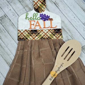 May include: A brown kitchen towel with a plaid hanging loop and embroidered text that says "hello fall" with a purple and orange leaf. A wooden spoon with the text "1B Embroidery Designs" is lying on top of the towel.