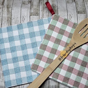 May include: Two gingham patterned pot holders in blue and pink/green with white accents. A wooden slotted spoon with the text "LB Embroidery Designs" rests on top. The pot holders have hanging loops.