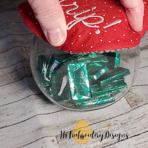 May include: A red fabric lid with white embroidery that says "Oh, a gift!" is being placed on a clear glass jar filled with green mint sweets.