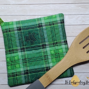 Pode incluir: Um pegador verde xadrez com acolchoado de trevo e uma argola de fita de bolinhas verdes e brancas. Uma colher de pau com fendas repousa ao lado do pegador. O pegador é quadrado e tem um desenho de trevo acolchoado.
