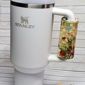 May include: White Stanley travel mug with a floral fabric handle wrap. The mug has a stainless steel lid and a logo of a bear with a crown on its head. The text "STANLEY" is printed below the bear.