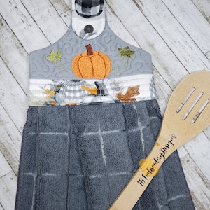Puede incluir: Una toalla de cocina a cuadros gris y blanco con una calabaza y hojas bordadas en ella. La toalla tiene un lazo para colgar y está unida a una cuchara de madera con el texto "1B Embroidery Designs" escrito en ella.