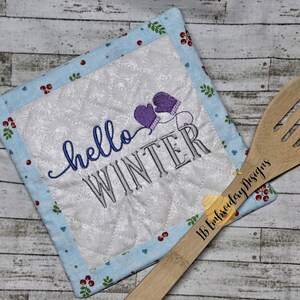 May include: A blue and white quilted pot holder with the text "hello winter" and a pair of purple mittens embroidered on it. The pot holder is lying on a wooden surface with a wooden spoon with the text "1B Embroidery Designs" on it.