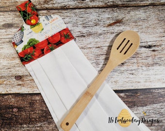 May include: White kitchen towel with a red and green tomato print. The towel has a hanging loop and is folded in thirds. A wooden spoon is lying on top of the towel. The towel has the text "Garden Fresh" and "Italy in a Jar" printed on it. The text "LB Embroidery Designs" is printed in the bottom right corner of the towel.