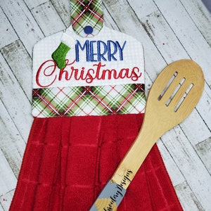 May include: Red and white plaid kitchen towel with a green and white stocking and the words "Merry Christmas" embroidered on it. The towel is hanging on a wooden spoon.