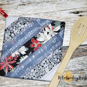 May include: A grey and white quilted pot holder with a Christmas theme. The fabric features musical notes, holly, berries, and a floral pattern. The pot holder has a red loop for hanging. "Merry Christmas" is written on the loop.