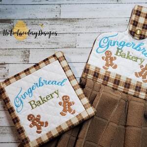 May include: A set of two kitchen towels with a gingerbread theme. The towels are brown and white with a plaid pattern. The text "Gingerbread Bakery" is embroidered on both towels, along with gingerbread men.