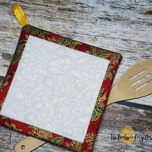 Può includere: Un sottopentola trapuntato rosso e verde con un centro bianco e ricami dorati. Il sottopentola ha un anello giallo per appenderlo. Il sottopentola è su una superficie di legno.