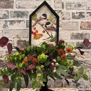 May include: A decorative centerpiece featuring a framed artwork of two birds perched on a branch with red berries. The artwork is set above a lush arrangement of green and burgundy foliage, accented with orange and cream-colored accents. The base is a black pedestal.