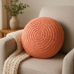 Puede incluir: Un cojín redondo de punto de color coral con un patrón circular. El cojín está sentado en un sofá beige claro.